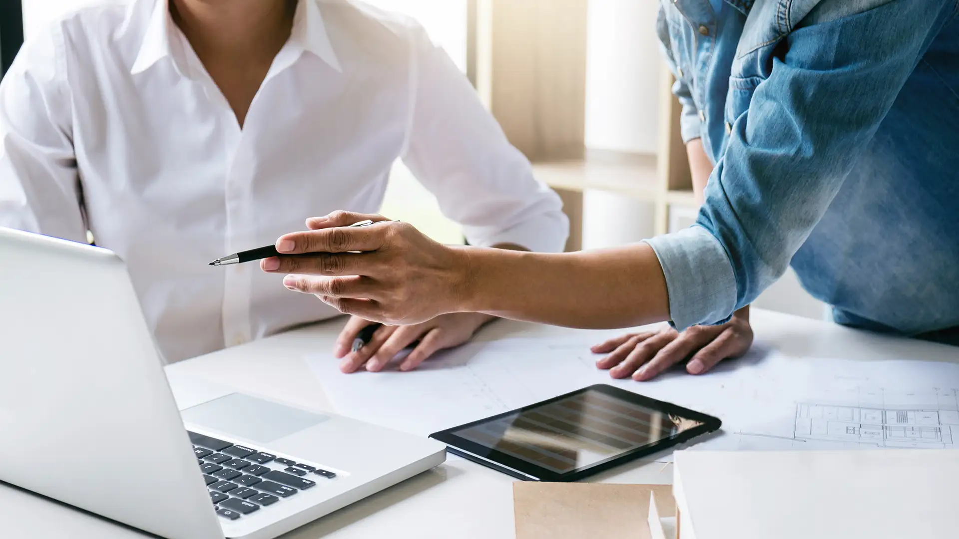 Två personer samarbetar vid ett skrivbord med laptop, ritningar och surfplatta.